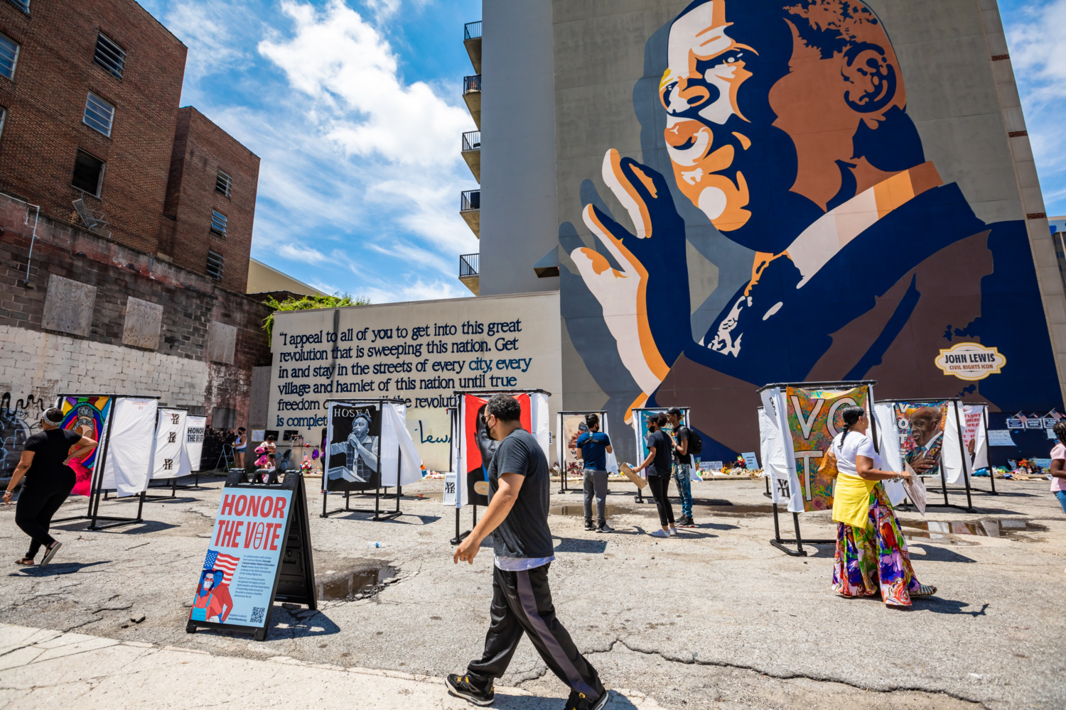 Photo of an art activation featuring voting booths each with a different mural painted onto it highlighting voting rights issues and civil rights icons. In the background there is a large mural of John Lewis.