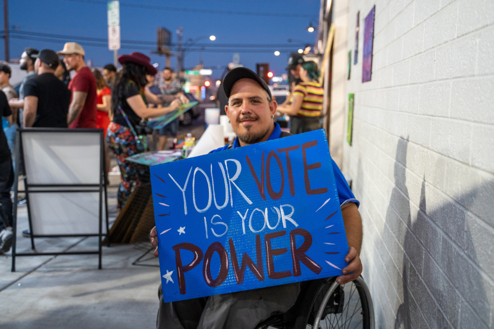 Person using wheelchair holds up a sign that reads "Your Vote Is Your Power" at an event