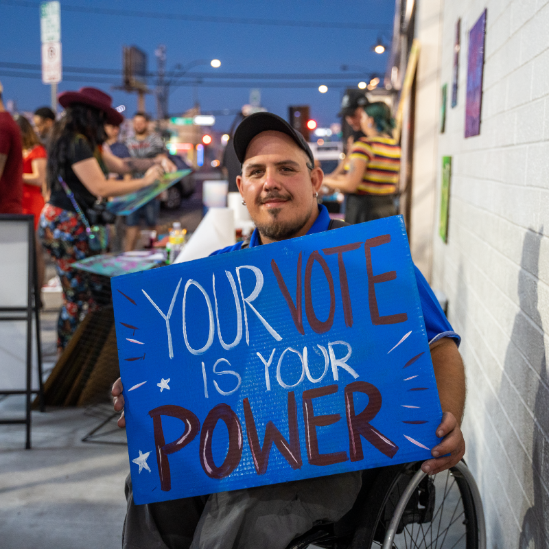 Person using wheelchair holds up a sign that reads "Your Vote Is Your Power" at an event