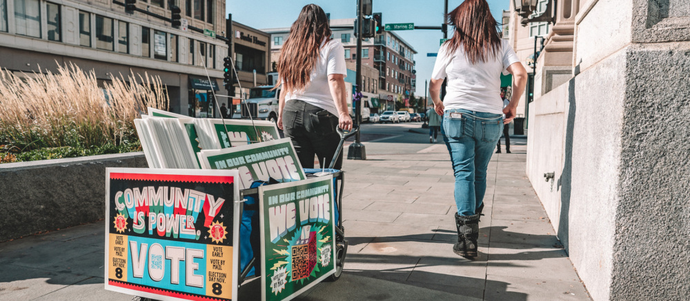 Two people pulling a cart of signs encouraging people to vote, for the sake of their community.