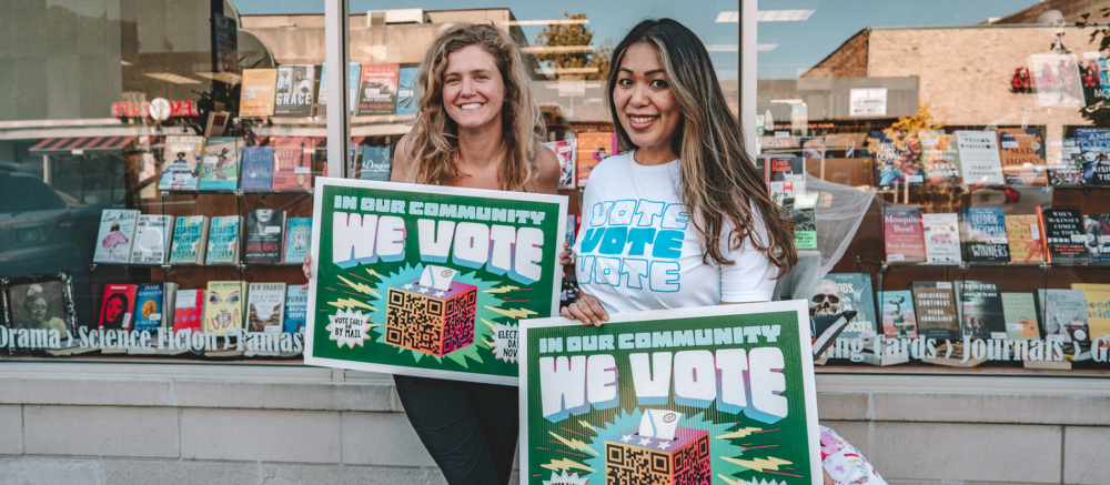 Two people standing in front of a bookstore holding signs that say "In Our Community, We Vote".