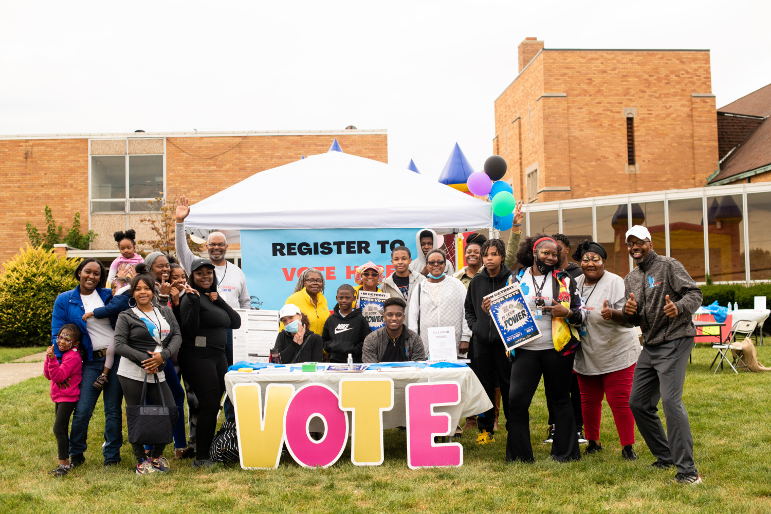 A group poses for a photo at a Democracy For All Event, signs include "Vote" and "Register to vote here"