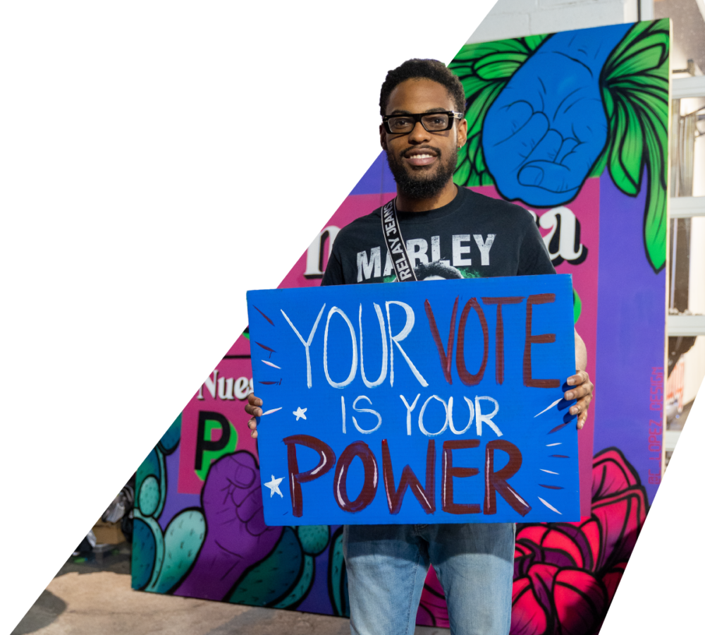 Man holds "Your Vote Is Your Power" Sign at community event in front of mural