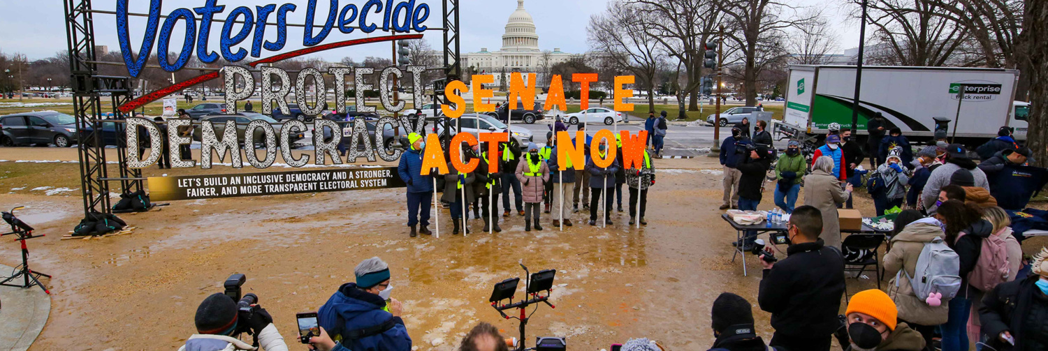 Activists holding signs that spell out "Senate Act Now" in front of an art installation about protecting democracy.