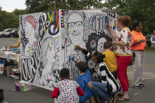 Members of the community fill in a mural with markers at a Civic Engagement Event. The mural reads "Our Vote is Our Power" and "Community is Everything"