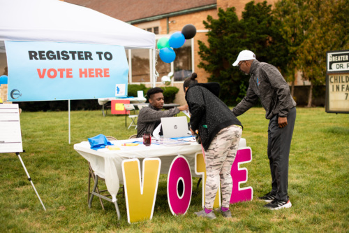 A volunteer helps two members of the community at a table with a sign saying "Register to Vote Here" at an outdoor community event