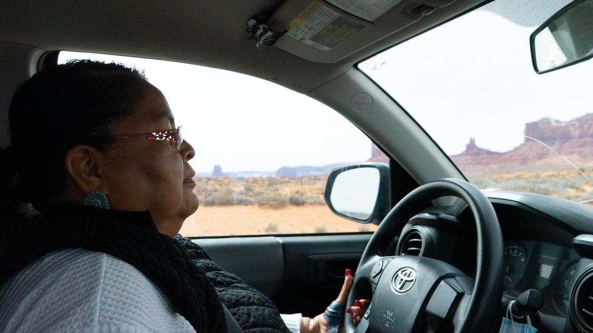 Side profile of a woman driving through a Utah desert with buttes in the distance, in a photo taken from the passenger's seat.