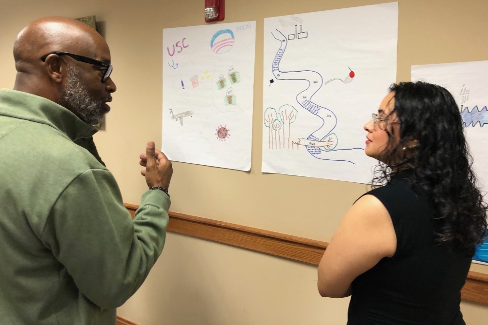 A man and woman stand discussing drawings depicting their leadership journeys taped to the wall in front of them.